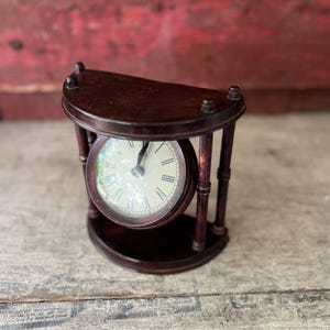 May include: A dark brown wooden clock with a cream-colored face and Roman numerals. The clock is encased in a semi-circular wooden frame with vertical supports. The hands point to approximately 1:05. The clock is a decorative item.