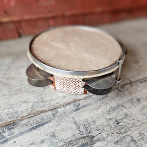 May include: A close-up of a tambourine with a light brown drumhead and a silver metal rim. The tambourine has small, silver jingles and a patterned band with white and brown geometric designs. The background is a weathered wooden surface.