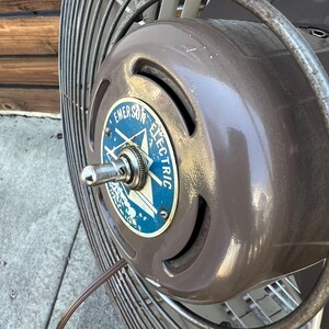 Pu&ograve; includere: Primo piano di un ventilatore vintage Emerson Electric. Il ventilatore ha un alloggiamento motore marrone con un emblema blu e bianco. Le pale del ventilatore sono racchiuse in una gabbia di metallo. Il ventilatore &egrave; un'antiquariato.