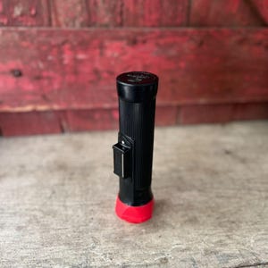 May include: A black flashlight with a red base and a black switch. The flashlight has a ribbed texture and a black top. The background is a weathered red wooden surface.