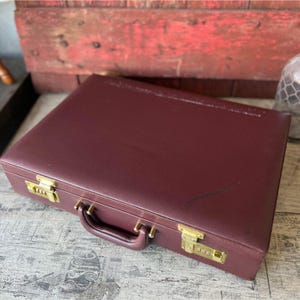 May include: A burgundy briefcase with gold-colored combination locks and handle. The briefcase is closed and appears to be made of leather or a leather-like material. The locks have three dials each.