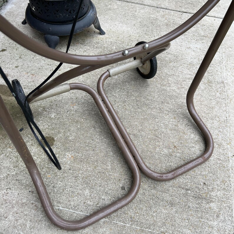 May include: A brown metal outdoor cart with two small wheels and a curved frame. The cart has a simple design and appears to be made of metal tubing. The cart is on a concrete surface.