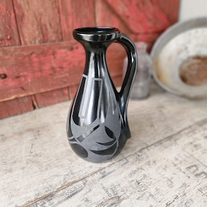 May include: A black ceramic pitcher with a handle and a decorative gray design. The pitcher has a narrow neck and a rounded body. The gray design features geometric and abstract shapes. The pitcher is on a light-colored wooden surface.