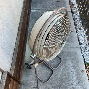 Pu&ograve; includere: Un ventilatore oscillante vintage beige e marrone con una gabbia circolare e una maniglia curva. Il ventilatore ha un supporto in metallo con ruote per un facile spostamento. Il ventilatore &egrave; posizionato su una superficie di cemento.