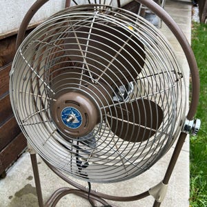 May include: A vintage Emerson Electric oscillating fan. The fan has a brown metal frame and a circular cage with a brown fan blade. The fan's motor housing is brown with a blue and white Emerson Electric logo. The fan is standing on a brown metal stand.