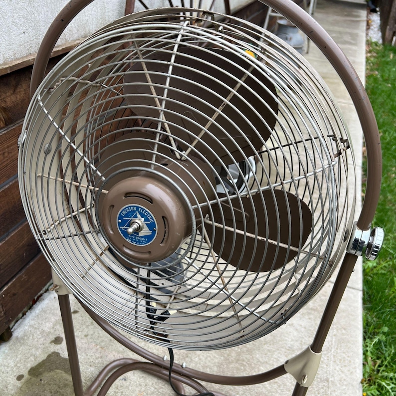 May include: A vintage Emerson Electric oscillating fan. The fan has a brown metal frame and a circular cage with a brown fan blade. The fan's motor housing is brown with a blue and white Emerson Electric logo. The fan is standing on a brown metal stand.