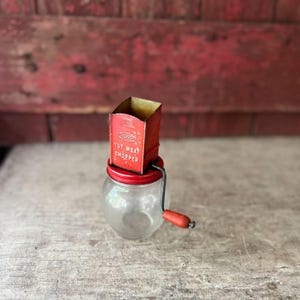 May include: A vintage Androck nut meat chopper featuring a red metal hopper with white text, a clear glass jar base, and a manual hand crank with a red wooden handle. The metal top is square-shaped and sits securely on the round glass container.
