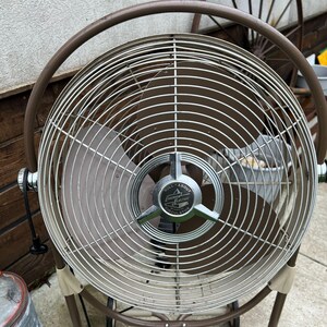 May include: A vintage Emerson Electric Roll-About Circulator fan. The fan has a brown metal frame and a silver-colored cage. The fan blades are a light brown color. The fan has a circular design and is standing on a concrete surface.