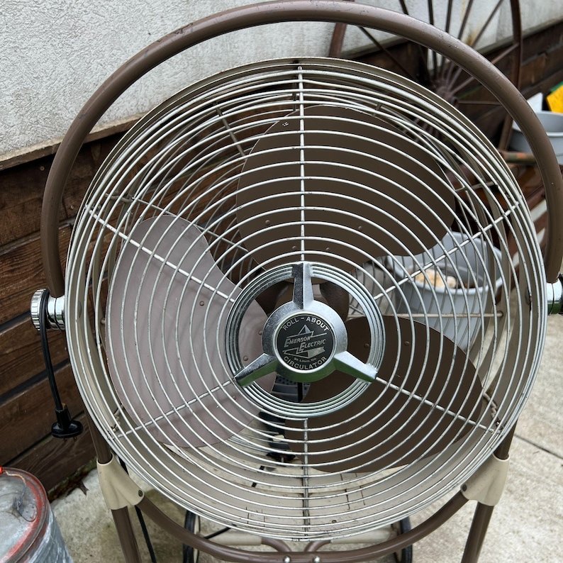 May include: A vintage Emerson Electric Roll-About Circulator fan. The fan has a brown metal frame and a silver-colored cage. The fan blades are a light brown color. The fan has a circular design and is standing on a concrete surface.