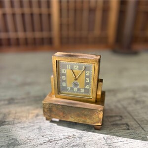 May include: A vintage gold-toned desk clock with a square face and a rectangular base. The clock face is a light gold color with black numerals and hands. The clock has a classic, art deco design.
