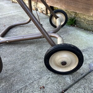 Pu&ograve; includere: Primo piano di un telaio di carrello in metallo marrone vintage con due ruote. Le ruote hanno pneumatici neri e centri biancastri. Il telaio del carrello &egrave; realizzato con tubi metallici curvi. Il carrello &egrave; su una superficie di cemento.