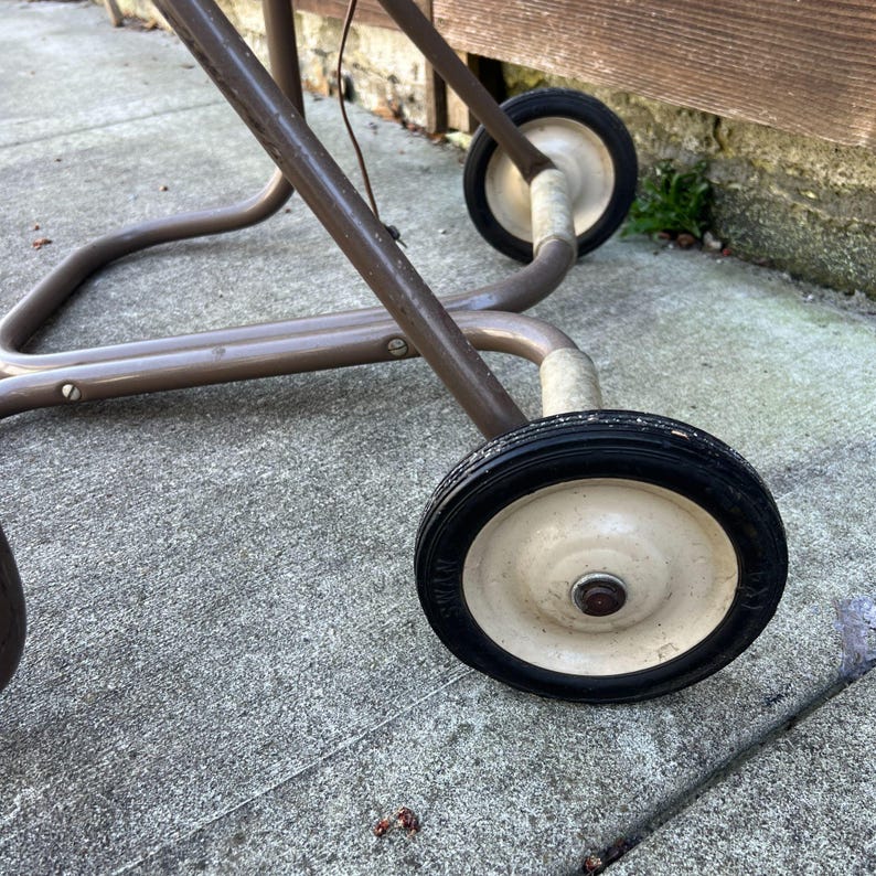 Pu&ograve; includere: Primo piano di un telaio di carrello in metallo marrone vintage con due ruote. Le ruote hanno pneumatici neri e centri biancastri. Il telaio del carrello &egrave; realizzato con tubi metallici curvi. Il carrello &egrave; su una superficie di cemento.