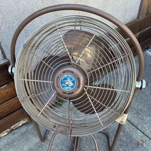 Pu&ograve; includere: Un ventilatore elettrico vintage marrone e argento con una gabbia protettiva e un design circolare. Il ventilatore ha tre pale e una base marrone. Il centro del ventilatore ha un emblema blu e bianco con la scritta "American Electric".