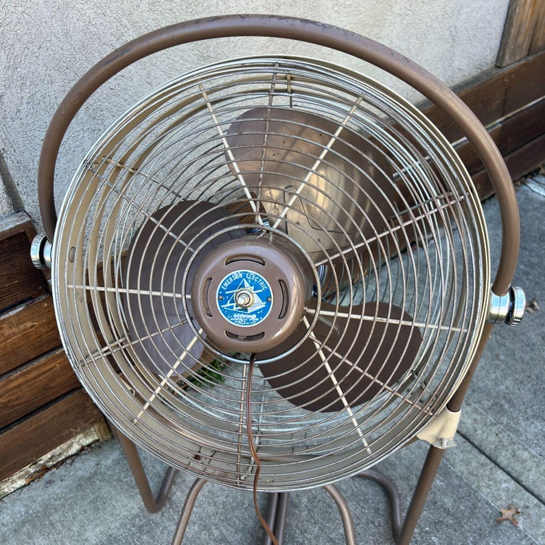 Pu&ograve; includere: Un ventilatore elettrico vintage marrone e argento con una gabbia protettiva e un design circolare. Il ventilatore ha tre pale e una base marrone. Il centro del ventilatore ha un emblema blu e bianco con la scritta "American Electric".