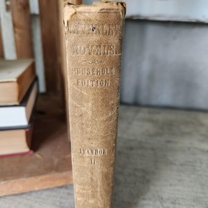 May include: A vintage book with a brown leather cover. The title is "The Works of Sir Walter Scott" in a gold font. The subtitle is "Household Edition" in a smaller gold font. The book is titled "Ivanhoe" in a black font.