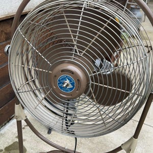May include: A vintage Emerson Electric oscillating fan with a brown metal frame and a circular cage. The fan has a brown motor housing with a blue and white emblem. The fan's blades are visible behind the metal cage.