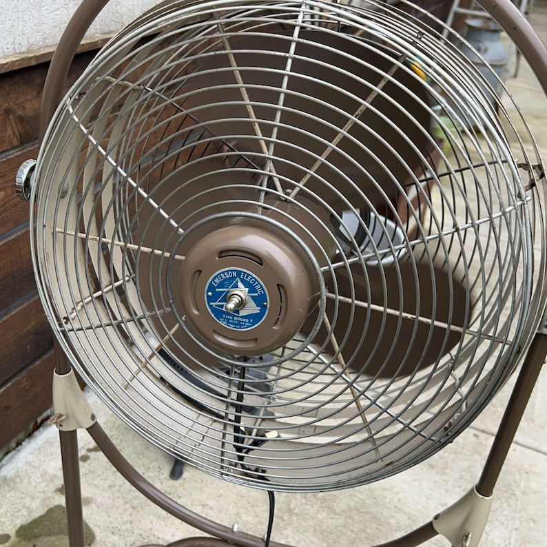 May include: A vintage Emerson Electric oscillating fan with a brown metal frame and a circular cage. The fan has a brown motor housing with a blue and white emblem. The fan's blades are visible behind the metal cage.