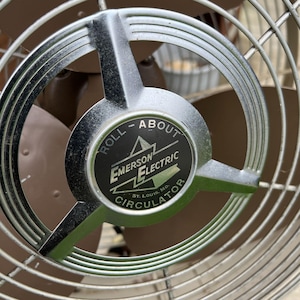 May include: Close-up of an Emerson Electric Circulator fan. The fan has a silver metal frame and a brown fan blade. The center of the fan has a black and white label that reads "ROLL - ABOUT" and "EMERSON ELECTRIC CIRCULATOR".