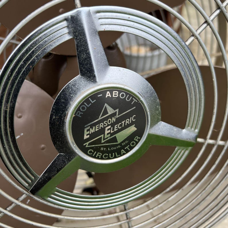 May include: Close-up of an Emerson Electric Circulator fan. The fan has a silver metal frame and a brown fan blade. The center of the fan has a black and white label that reads "ROLL - ABOUT" and "EMERSON ELECTRIC CIRCULATOR".