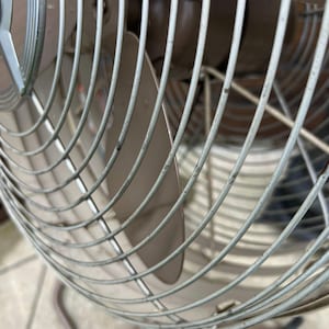 May include: Close-up of a vintage oscillating fan. The fan has a metal cage with curved, light gray bars. The blades are a light brown color. The fan is designed to oscillate and provide air circulation.