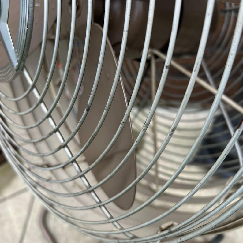May include: Close-up of a vintage oscillating fan. The fan has a metal cage with curved, light gray bars. The blades are a light brown color. The fan is designed to oscillate and provide air circulation.