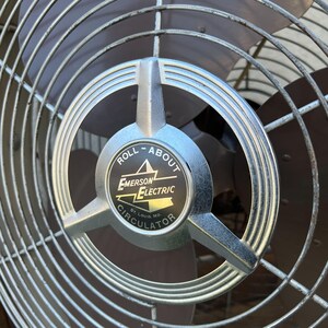Pu&ograve; includere: Primo piano di un ventilatore vintage Emerson Electric Roll-About Circulator. Il ventilatore presenta una gabbia in metallo argentato e un emblema centrale con il nome del marchio e le parole "Roll-About Circulator". Le pale sono marrone scuro.