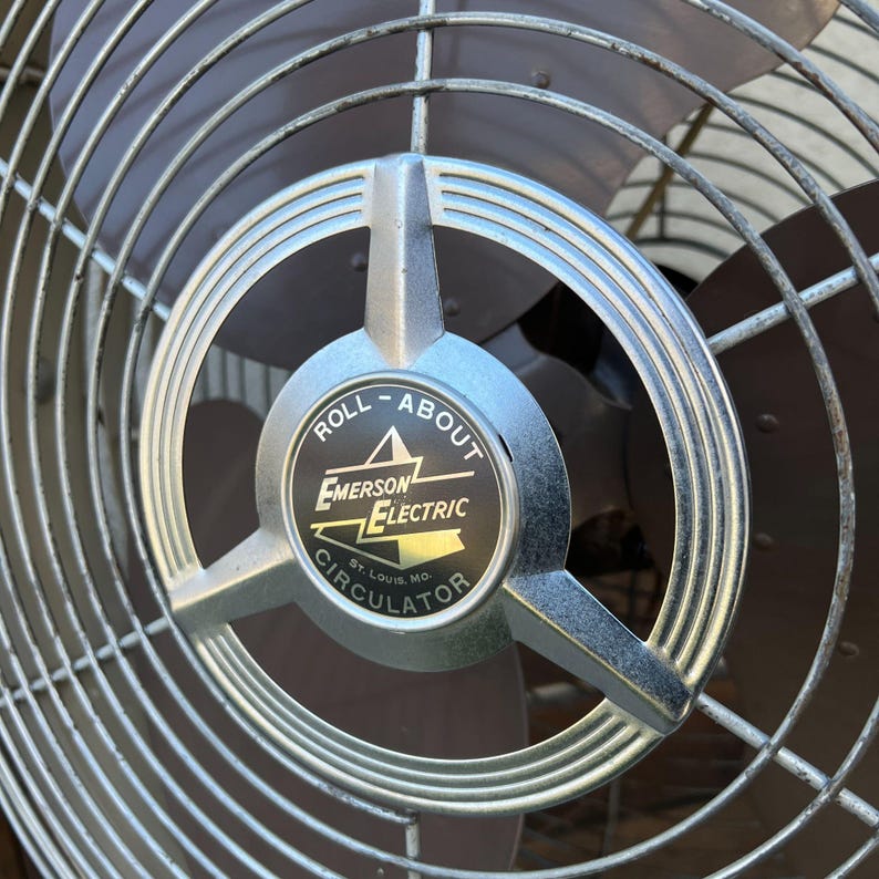 Pu&ograve; includere: Primo piano di un ventilatore vintage Emerson Electric Roll-About Circulator. Il ventilatore presenta una gabbia in metallo argentato e un emblema centrale con il nome del marchio e le parole "Roll-About Circulator". Le pale sono marrone scuro.