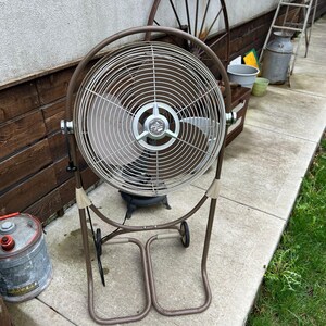 May include: A vintage brown and silver oscillating fan with a circular cage and a sturdy base with wheels. The fan has a classic design, perfect for cooling a room or outdoor space. The fan is made of metal and has a retro aesthetic.