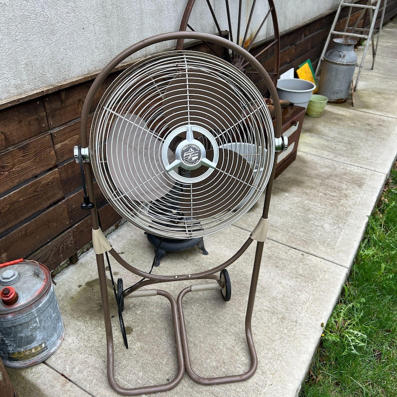 May include: A vintage brown and silver oscillating fan with a circular cage and a sturdy base with wheels. The fan has a classic design, perfect for cooling a room or outdoor space. The fan is made of metal and has a retro aesthetic.