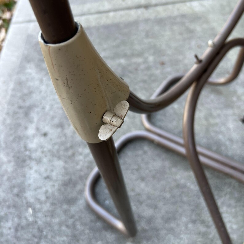Pu&ograve; includere: Primo piano di una sezione di un telaio di sedia in metallo. Il telaio &egrave; di colore marrone scuro, con un giunto beige e una vite a forma di farfalla. Sono visibili le gambe e i supporti curvi della sedia.