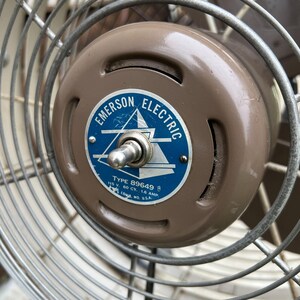 May include: Close-up of an Emerson Electric fan. The fan's motor housing is a muted brown, with a blue and white label that reads "Emerson Electric" and "Type 89649 B". The fan's cage is made of silver metal.