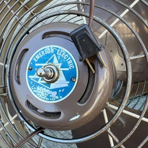 Pu&ograve; includere: Primo piano di un ventilatore vintage Emerson Electric. Il ventilatore ha un alloggiamento motore marrone con un logo blu e bianco. Le pale del ventilatore sono racchiuse in una gabbia di metallo. Il cavo di alimentazione e la spina sono visibili.