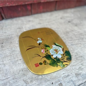 May include: A gold-colored, oval-shaped decorative plate with a floral design. The plate features white and pink flowers with green leaves and a small bird. There is also some text and a red stamp.