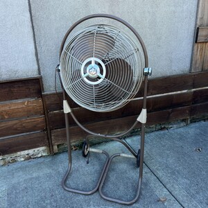 Pu&ograve; includere: Un ventilatore oscillante vintage in metallo marrone con una gabbia circolare e un supporto con ruote. La ventola ha una pala argentata e un emblema decorativo al centro. Il supporto ha un design curvo e un accento beige.