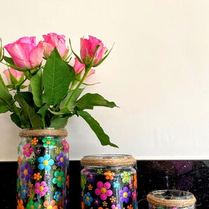 May include: Three glass jars with a floral design painted on them. The jars are different sizes and are decorated with brown twine around the top. The jars are filled with pink roses.