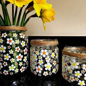 May include: Three glass jars with a rustic twine accent around the top. The jars are decorated with white and orange daisy flowers. The largest jar is filled with yellow daffodils.