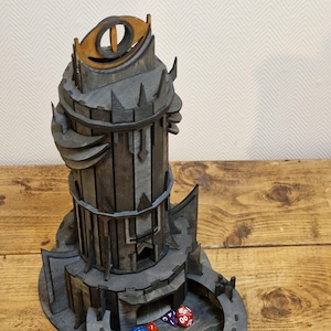 May include: A wooden tower-shaped dice rolling tower with a dark gray finish and a golden eye symbol on top. The tower has multiple levels and a circular dice tray at the bottom. There are several colorful dice scattered around the tower.