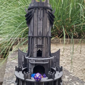 May include: A dark gray wooden tower with a black eye symbol on top, designed as a dice rolling tower for tabletop games. The tower has a circular base with a moat and a central chamber for dice to fall into. Several colorful dice are visible inside the tower and on the base.