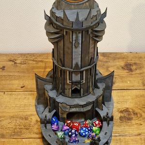 May include: A wooden tower-shaped dice rolling tower with a large eye symbol on top. The tower is painted in a dark gray color and has a compartment at the bottom to hold dice. The compartment is filled with colorful dice.