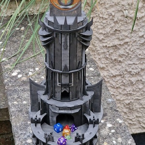 May include: A black wooden tower with a large eye on top, designed for rolling dice. The tower has a central chamber where dice can be rolled and collected. There are several colorful dice scattered around the base of the tower.