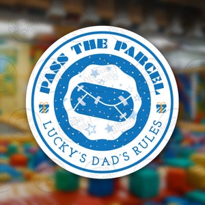 May include: A round sticker with a blue and white design. The text "PASS THE PARCEL" and "LUCKY'S DAD'S RULES" are in a blue ring around a blue parcel graphic. Small gift box graphics and star shapes are also included.