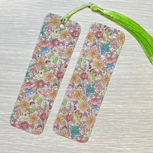 May include: Two rectangular bookmarks with a floral and character design. The design includes flowers, mushrooms, and cartoon characters in shades of red, yellow, green, and blue. Each bookmark has a green tassel attached at the top.