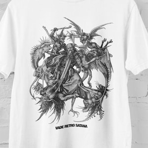 May include: White t-shirt featuring a detailed black and white illustration of a chaotic scene with figures, demons, and creatures. The text "VADE RETRO SATANA" is printed below the graphic. The shirt is hanging on a wooden hanger against a white brick wall.