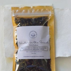 May include: A clear plastic bag filled with loose leaf tea, labeled "Winter in the Forest Herbal Tea Blend." The label lists ingredients like black tea, holy basil, and hibiscus. The bag is set on a white surface.