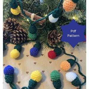 May include: A string of crocheted light bulbs in various colors, including red, yellow, blue, green, orange, and white. The bulbs are connected by green yarn and are arranged on a brown surface with a heart pattern. The text "Pdf Pattern" is visible in the upper right corner of the image.