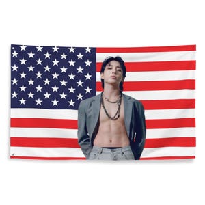 Puede incluir: Una bandera estadounidense rectangular con rayas rojas y blancas y un campo azul con estrellas blancas. Una persona está de pie frente a la bandera, vistiendo una blazer gris, vaqueros y collares.