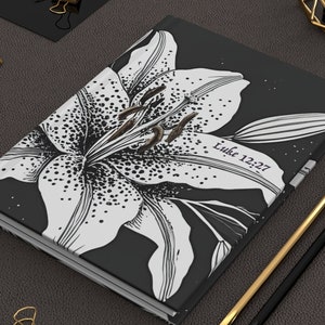 May include: A black journal with a white lily flower design and the text "Luke 12:27". The journal is next to a gold and a black pencil, and gold binder clips.