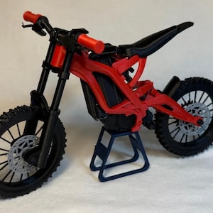 May include: A red and black miniature dirt bike model with a black seat, tires, and suspension. The handlebars have red grips. The bike is displayed on a small blue stand. The wheels have a silver-colored disc brake.