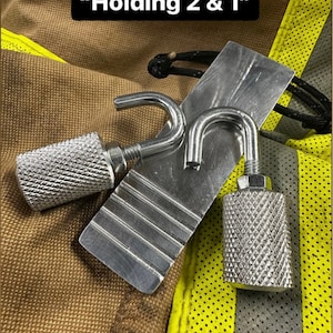 May include: Two silver hooks with a silver metal plate with a textured surface. The plate has four grooves. The text "Holding 2 & 1" is visible above the hooks. The hooks are on a brown and yellow fabric background.