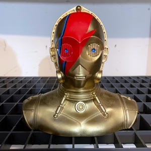 Puede incluir: Un busto dorado de C-3PO con un diseño de rayo rojo y azul en la cara. El busto tiene un acabado metálico y se exhibe en una estantería de rejilla negra. Los ojos son azules.